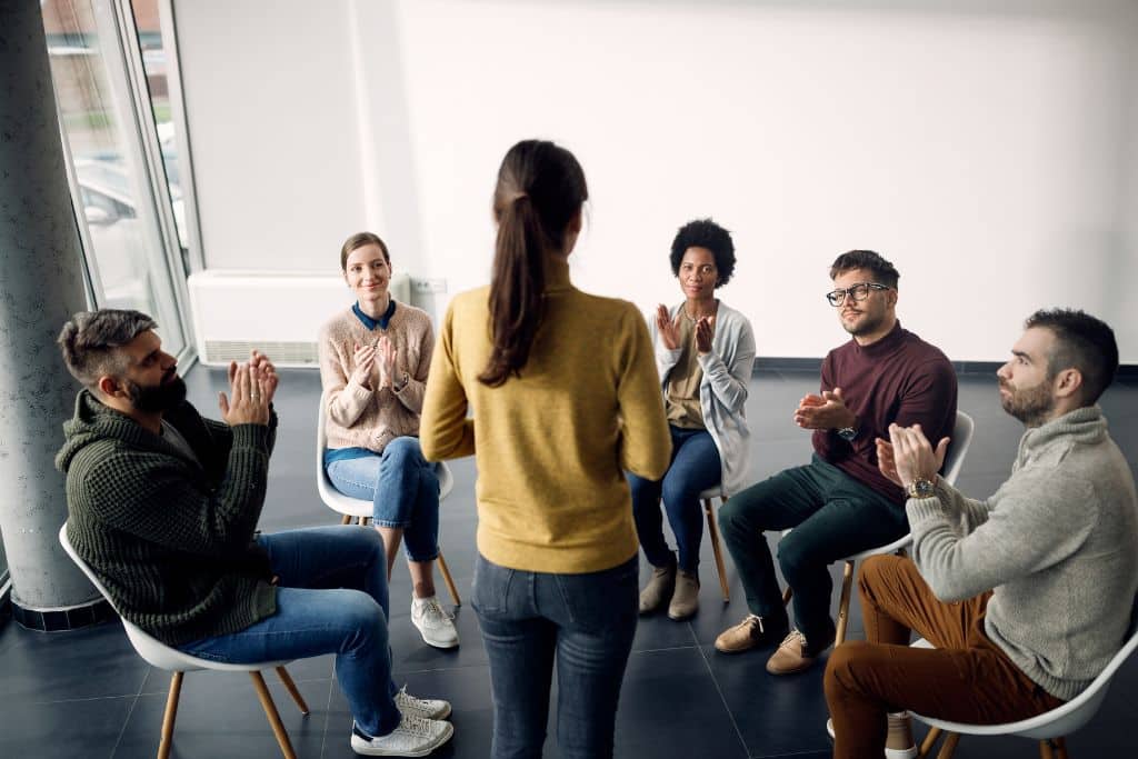 Alcohol addiction group therapy attenders applauding one participant during meeting | Which Rehab