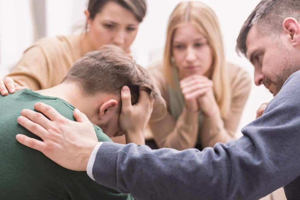 A group of people supporting a man with his head in his hands at a group addiction therapy session - Which Rehab