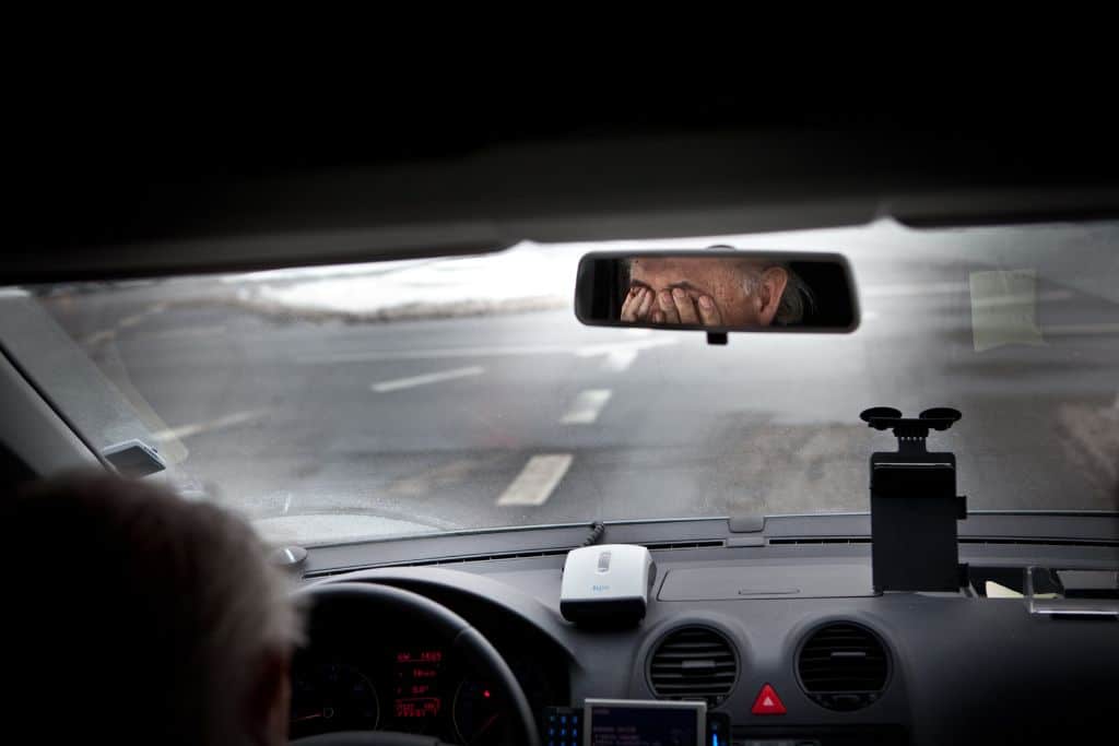 A rear view mirror view of a sad man sitting in a car rubbing his eyes - Which Rehab