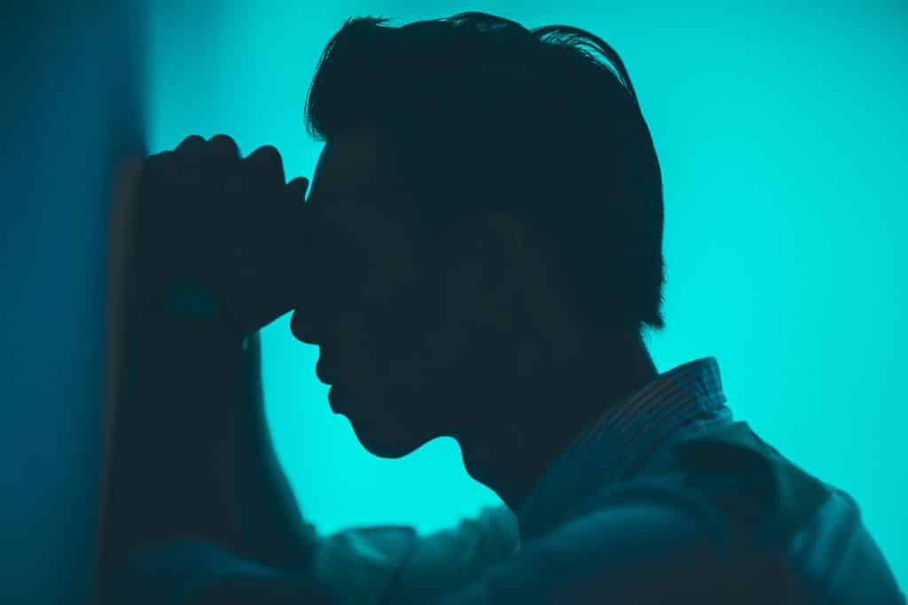 A silhouette of a man in a blue lit room with his head against the wall - Which Rehab